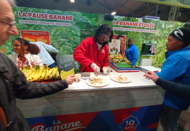 Sur le stand d'un salon, une personne prend une rondelle de banane pour dégustation, qu'un homme en arrière plan vient de couper
