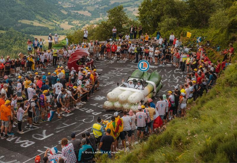 photo prise sur l'étape 11 du tour de france 2024 : la voiture en forme de poireau de E.Leclerc est saluée par la foule. Derrière vient la moto en forme de cerise.