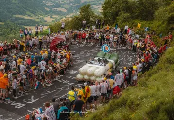 photo prise sur l'étape 11 du tour de france 2024 : la voiture en forme de poireau de E.Leclerc est saluée par la foule. Derrière vient la moto en forme de cerise.