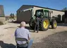 Un homme plutôt âgé assis sur une chaise dans une cour de ferme regardant un agriculteur devant son tracteur.
