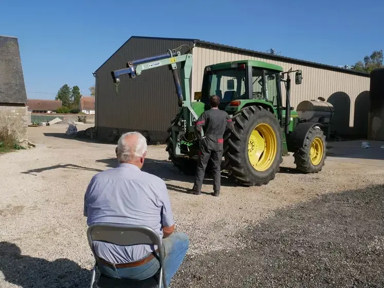 Un homme plutôt âgé assis sur une chaise dans une cour de ferme regardant un agriculteur devant son tracteur.