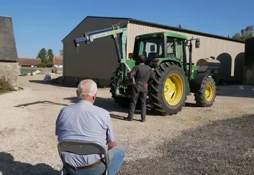Un homme plutôt âgé assis sur une chaise dans une cour de ferme regardant un agriculteur devant son tracteur.