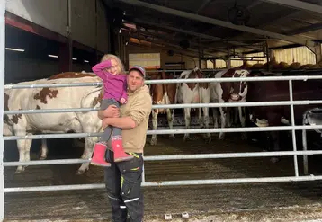 Un éleveur avec sa petite fille dans ses bras devant ses vaches rentrées dans la stabulation.