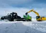 Chantier de transport de neige avec une dameuse tractant une remorque Joskin à chenilles à coté d'une pelle sur chenilles