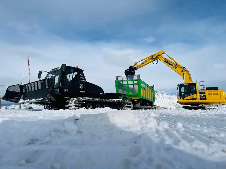 Chantier de transport de neige avec une dameuse tractant une remorque Joskin à chenilles à coté d'une pelle sur chenilles