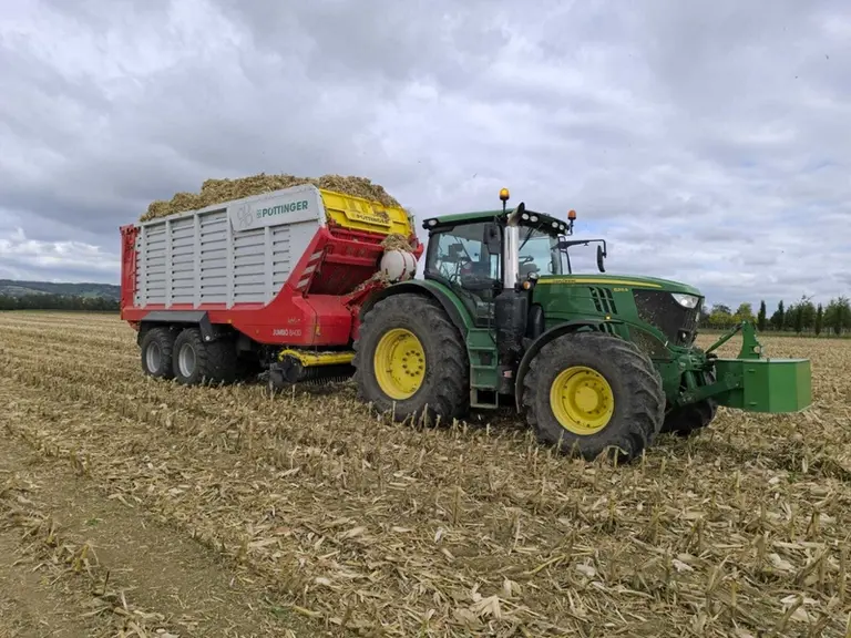 Tracteur John Deere 6R215 et remorque autochargeuse Pöttinger Jumbo 8400 à la récolte de paille de maïs.