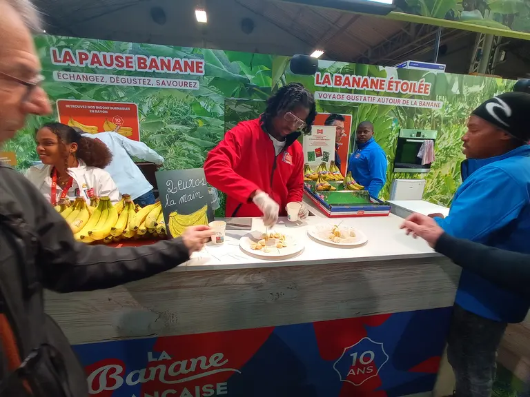 Sur le stand d'un salon, une personne prend une rondelle de banane pour dégustation, qu'un homme en arrière plan vient de couper