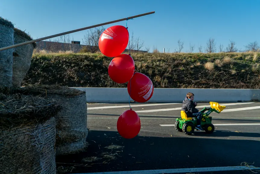 Des ballons rouges à l'effigie des jeunes agriculteurs accrochés au bout d'un bâton planté dans des bottes de paille. En arrière plan, un enfant sur u