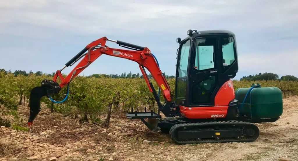 Mini-pelle Kubota dans les vignes équipée d'un godet planteur et d'une cuve d'arrosage.