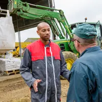 Discussion sur la ferme entre un agriculteur et son salarié, big bag et tracteur en arrière plan