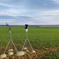 Station météo Sencrop avec anémomètre et pluviomètre devant une parcelle de céréales.