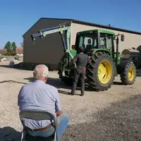 Un homme plutôt âgé assis sur une chaise dans une cour de ferme regardant un agriculteur devant son tracteur.