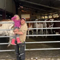 Un éleveur avec sa petite fille dans ses bras devant ses vaches rentrées dans la stabulation.