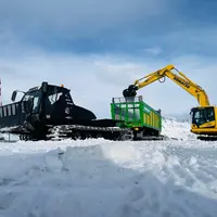 Chantier de transport de neige avec une dameuse tractant une remorque Joskin à chenilles à coté d'une pelle sur chenilles