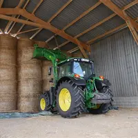 Tracteur John Deere avec chargeur devant un stockage de balles de paille rondes.