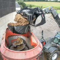 Benne à grappin coupe-balle Xtra’Shark de Magsi chargeant une balle d'enrubannage dans une remorque mélangeuse à vis vert
