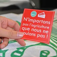 Une main tient un sticker avec écrit "n'importons pas l'agriculture que nous ne voulons pas"