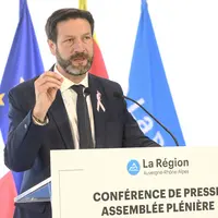 un homme de 50 ans parlant devant une tribune, derrière lui, des drapeaux