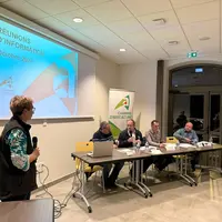 Une femme se tient debout avec un micro à côté d'un écran. En arrière plan 4 hommes sont assis à une table, l'un d'entre eux explique quelque chose au