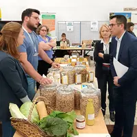 Dans une salle, au milieu d'autres personnes, deux hommes debout parlent autour d'une table avec des produits agricoles