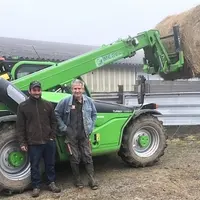 Deux hommes souriants, un jeune et un plus âgé, sont appuyés sur une machine agricole.