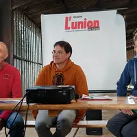 Trois hommes, à une tribune improvisée dans une stabulation agricole, sourient.