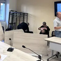 Tables renversées dans une salle de réunion, avec Christian Arvis s'adressant à une personne hors cadre avec une véhémence gestuelle visible.