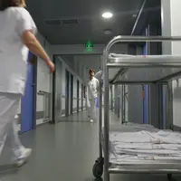 Couloir d'hôpital avec du personnel soignant en blouse blanche qui marche. Un brancard avec des blouse est sur le premier plan à droite de l'image.