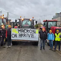 Agriculteurs de la Coordination rurale portant des bonnets jaunes devant des tracteurs dans un village avec une pancarte accrochée sur un tracteur sur