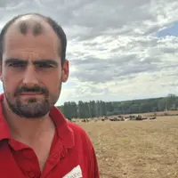 Un jeune éleveur devant sa prairie jaunie avec ses génisses au fond