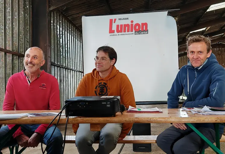 Trois hommes, à une tribune improvisée dans une stabulation agricole, sourient. 