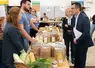 Dans une salle, au milieu d'autres personnes, deux hommes debout parlent autour d'une table avec des produits agricoles