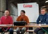 Trois hommes, à une tribune improvisée dans une stabulation agricole, sourient. 