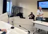 Tables renversées dans une salle de réunion, avec Christian Arvis s'adressant à une personne hors cadre avec une véhémence gestuelle visible.