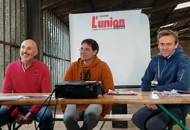 Trois hommes, à une tribune improvisée dans une stabulation agricole, sourient. 
