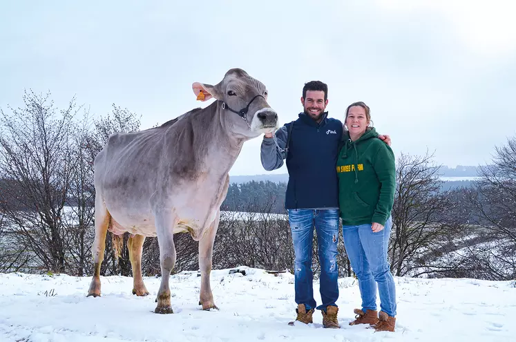 Un couple d'éleveurs souriant aux côtés de leur vache de race brune dans la neige.