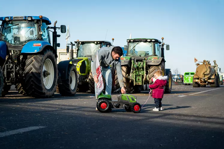 Un papa aide sa petite fille avec son tracteur à pédale sur l'A71 de Clermont-Ferrand, devant une file de tracteurs stationnés..