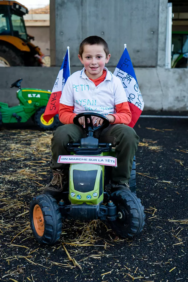 Petit garçon sur un tracteur à pédales avec deux drapeaux français et un panneau communal miniature retourné accroché à l'avant.