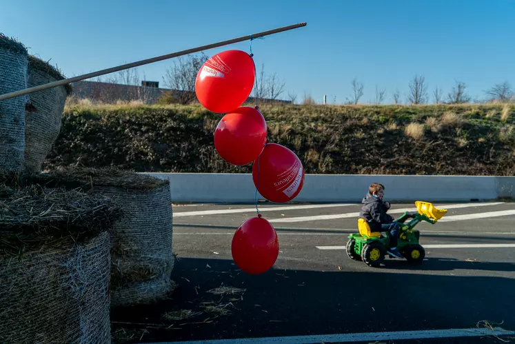 Des ballons rouges à l'effigie des jeunes agriculteurs accrochés au bout d'un bâton planté dans des bottes de paille. En arrière plan, un enfant sur un tracteur à pédales.