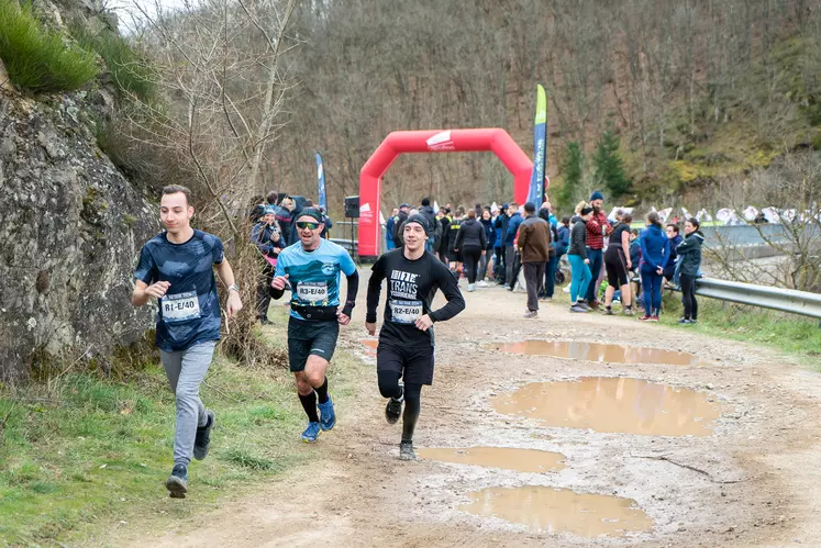 Trois coureurs côte à côte sur un chemin gadouilleux, un groupe de gens en arrière plan.