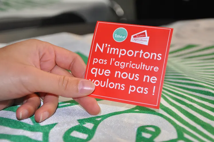 Une main tient un sticker avec écrit "n'importons pas l'agriculture que nous ne voulons pas"