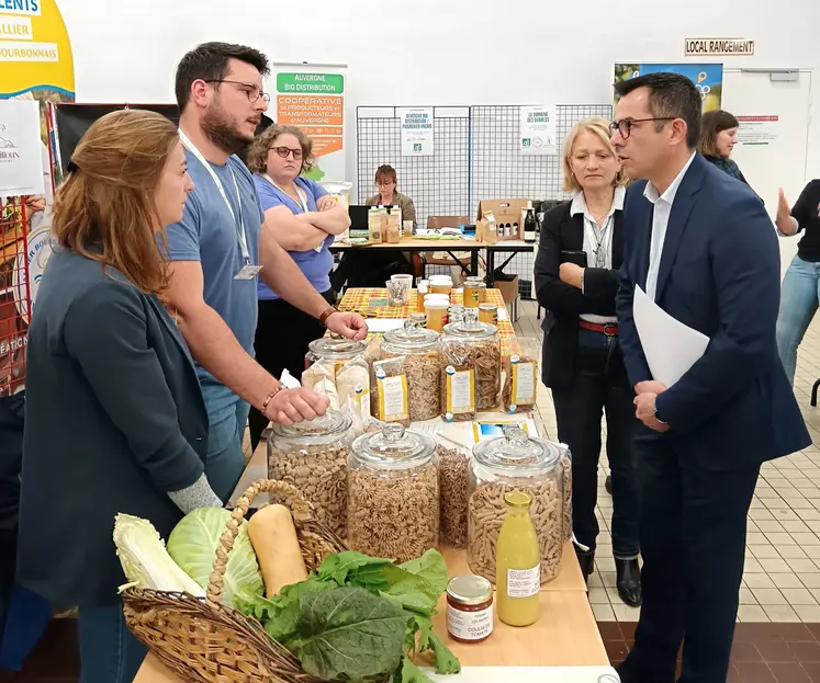 Dans une salle, au milieu d'autres personnes, deux hommes debout parlent autour d'une table avec des produits agricoles