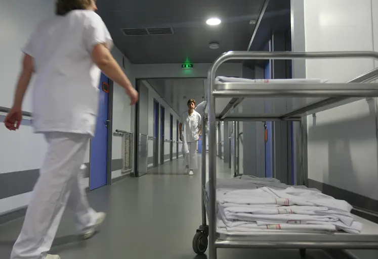 Couloir d'hôpital avec du personnel soignant en blouse blanche qui marche. Un brancard avec des blouse est sur le premier plan à droite de l'image.  