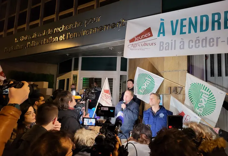 Mobilisations d'agriculteurs de la FRSEA et les JA d'Ile-de-France devant le siège de l'Inrae. Des journalistes filment un agriculteur, micro à la main. Des drapeaux des syndicats et un bout de banderole sont visibles. C'est le début de la journée. 