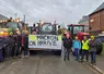 Agriculteurs de la Coordination rurale portant des bonnets jaunes devant des tracteurs dans un village avec une pancarte accrochée sur un tracteur sur laquelle on peut lire "Macron on arrive"