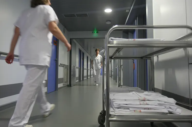 Couloir d'hôpital avec du personnel soignant en blouse blanche qui marche. Un brancard avec des blouse est sur le premier plan à droite de l'image.  