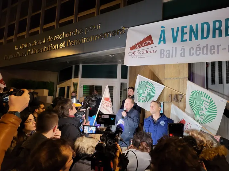 Mobilisations d'agriculteurs de la FRSEA et les JA d'Ile-de-France devant le siège de l'Inrae. Des journalistes filment un agriculteur, micro à la main. Des drapeaux des syndicats et un bout de banderole sont visibles. C'est le début de la journée. 