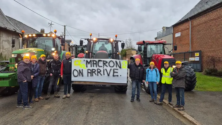 Agriculteurs de la Coordination rurale portant des bonnets jaunes devant des tracteurs dans un village avec une pancarte accrochée sur un tracteur sur laquelle on peut lire "Macron on arrive"