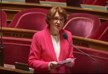 La nouvelle ministre de l'Agriculture Annie Genevard au Sénat, répondant dans un micro à un Sénateur. Mme Genevard porte une veste rose et tient des fiches dans ses mains. Les fauteuils rouge derrière elle sont vides. 