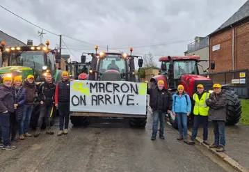 Agriculteurs de la Coordination rurale portant des bonnets jaunes devant des tracteurs dans un village avec une pancarte accrochée sur un tracteur sur laquelle on peut lire "Macron on arrive"